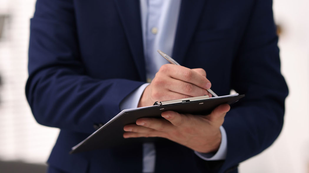 Personne vêtue d’un veston foncé écrivant sur un formulaire posé sur un porte‑documents